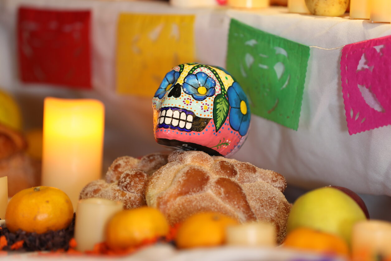 A small painted skull sits on top of a piece of bread on a traditional Mexican ofrenda.