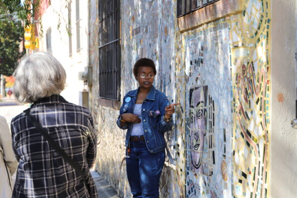 A woman tour guide gestures to a mosaic mural behind her.